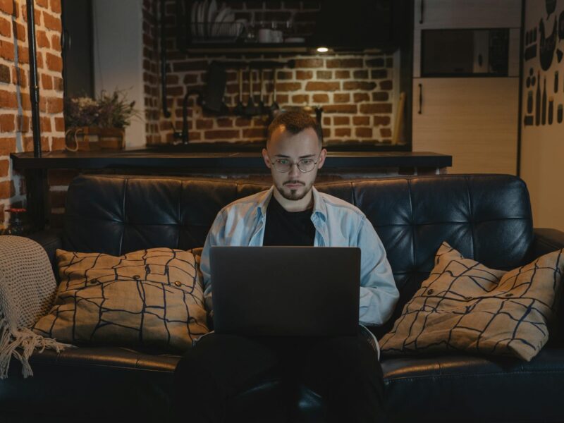 man in black crew neck t shirt and denim jacket sitting on a couch using his laptop